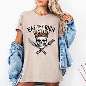May include: A beige t-shirt with the text "EAT THE RICH" printed in black. A skull wearing a gold crown is depicted with crossed cutlery.