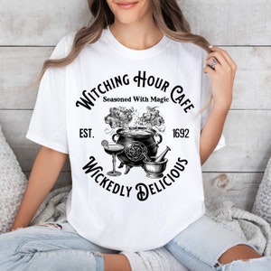 May include: White t-shirt featuring a black and white graphic design for the "Witching Hour Cafe." The design includes a cauldron, cocktail glass, mortar and pestle, and the text "Seasoned With Magic" and "Wickedly Delicious."