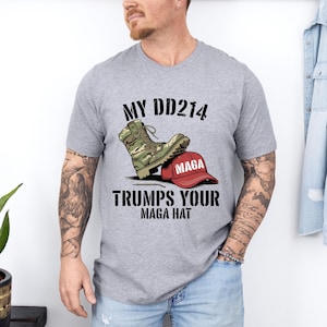Puede incluir: Una camiseta gris con el texto "My DD214 Trumps Your MAGA Hat" y un gráfico de una bota de camuflaje pisando un sombrero rojo "MAGA".