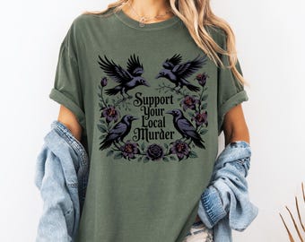 Puede incluir: Camiseta verde oliva con un diseño gráfico de cuatro cuervos negros, flores moradas y el texto "Support Your Local Murder". El diseño está centrado en la parte delantera de la camiseta.