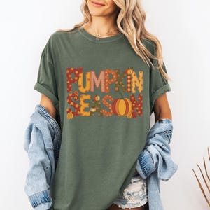 May include: Olive green t-shirt with the words "PUMPKIN SEASON" in a fall-themed design. The text features orange, yellow, and brown patterns and a pumpkin graphic. The shirt has rolled-up sleeves.