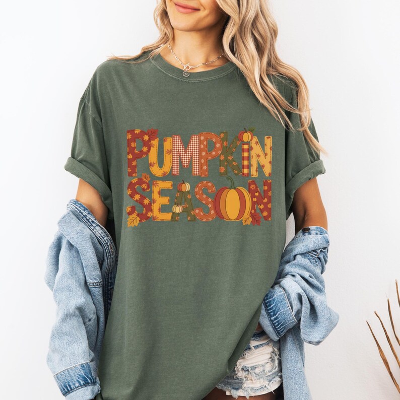 May include: Olive green t-shirt with the words "PUMPKIN SEASON" in a fall-themed design. The text features orange, yellow, and brown patterns and a pumpkin graphic. The shirt has rolled-up sleeves.