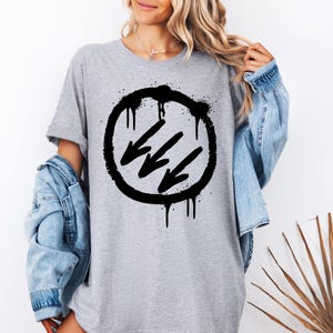 May include: A gray t-shirt featuring a black graphic design. The design includes a circle with a spray-paint effect, three downward-pointing arrows, and dripping paint details. The shirt has short sleeves and a relaxed fit.