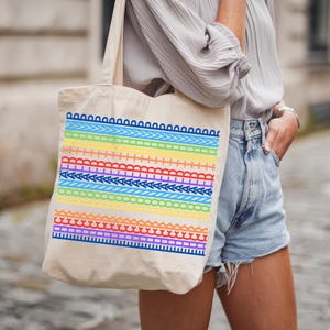 May include: Beige canvas tote bag featuring a colorful, rainbow-striped design. The design includes various geometric patterns in shades of red, orange, yellow, green, blue, and purple. The bag has long straps for carrying over the shoulder.