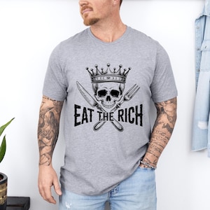 May include: Gray t-shirt with a black and white illustration of a skull wearing a crown and holding a knife and fork. The text "EAT THE RICH" is printed below the skull.