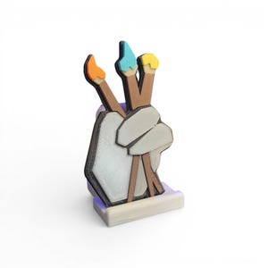 May include: A wooden art-themed figurine depicting a hand holding three paintbrushes. The paintbrushes have orange, blue, and yellow bristles. The hand is white with a black outline, and the base is light grey.