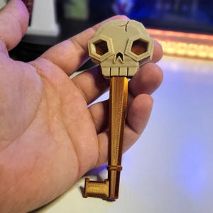Puede incluir: Una llave impresa en 3D con una cabeza en forma de cráneo. La llave está hecha de plástico beige y tiene un eje de metal dorado.