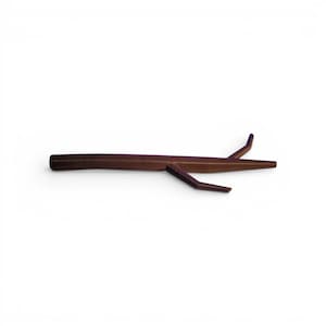 Puede incluir: Una pinza para el pelo en forma de ramita marrón, con dos pequeñas ramas que se extienden del cuerpo principal. La pinza está hecha de un material oscuro, posiblemente de madera, y está sobre un fondo blanco. El diseño es minimalista e inspirado en la naturaleza.