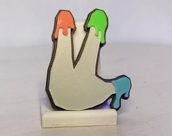 Puede incluir: Una letra L impresa en 3D de color blanco hueso con un borde marrón oscuro. La parte superior del trazo vertical tiene un efecto de goteo naranja, la parte superior del trazo diagonal un efecto verde brillante y la esquina inferior derecha azul.