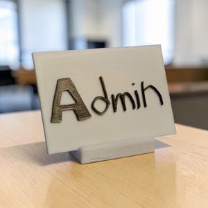 May include: A white rectangular desk sign featuring the word Admin in raised, dark, textured lettering. The sign is supported by a matching white base, creating a clear, upright display for office identification or workspace labeling.