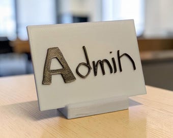 Puede incluir: Un letrero de escritorio rectangular blanco con la palabra Admin en letras oscuras, texturizadas y en relieve. El letrero se apoya en una base blanca a juego, creando una pantalla vertical clara para la identificación en la oficina.