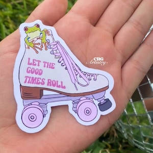 Pode incluir: Um patins branco com cadarços e rodas cor-de-rosa. Um sapo verde está sentado no topo do patins. O patins tem o texto "Let the good times roll" escrito no lado.