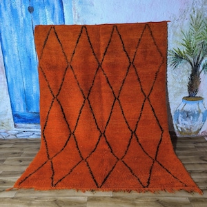 以下が含まれることがあります： 黒いひし形模様のオレンジ色のラグ。ラグは厚く、質感のある素材でできており、下端にフリンジが付いています。青いドアと鉢植えの植物のある壁を背景にラグが展示されています。