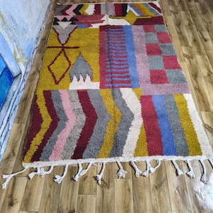 以下が含まれることがあります： 幾何学模様が施された長方形のカラフルなラグ。黄色、赤、青、ピンク、グレーのセクションがあります。白いタッセルが端を飾っています。ラグのデザインには、ダイヤモンド、ジグザグ、ストライプのパターンが含まれています。