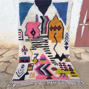May include: A colorful, geometric patterned rug with a cream-colored background. The rug features bold shapes in blue, pink, yellow, and black, with a fringed edge. The design includes triangles, zigzags, and other abstract forms.