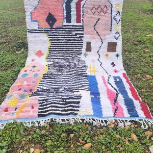 May include: A colorful, abstract rug with a white base and geometric shapes in pink, blue, black, and red. The rug features a striped pattern and fringe along the bottom edge. The rug is displayed outdoors on a grassy surface.
