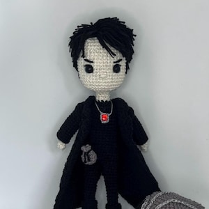 May include: A crocheted doll with black hair and a black outfit, holding a small black bag. The doll is standing next to a gray crocheted mask with a white crocheted tail and a small black crocheted bird.