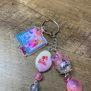 Puede incluir: Un llavero con temática de Barbie con un llavero y una cadena plateados. El llavero presenta un dije rectangular con el texto "IT'S GIRL'S NIGHT IN!" y un gráfico de Barbie. Los dijes adicionales incluyen un óvalo con el logotipo de Barbie, un oso rosa y un orbe rosa.