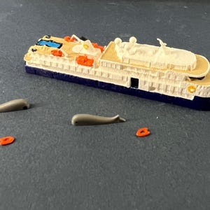 Op de afbeelding: Een wit en blauw speelgoed cruiseschip met een zwembad op het bovendek. Het schip is omringd door drie grijze speelgoedwalvissen en drie oranje reddingsboeien.