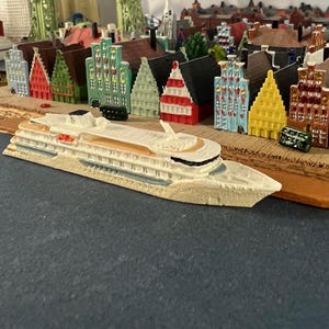 May include: A white cruise ship model with a brown deck and a gray hull. The ship is docked at a miniature harbor with colorful buildings.