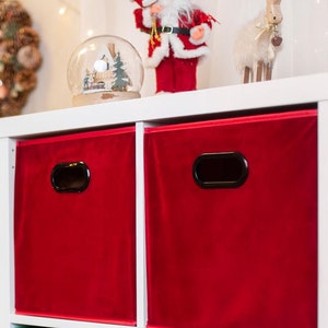 Puede incluir: Una estantería de almacenamiento de cubos blanca con seis cestas de almacenamiento de tela. Las cestas son rojas y verdes. Un árbol de Navidad con adornos dorados está en el fondo.