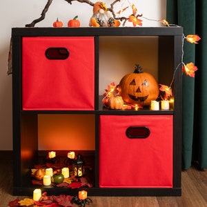 Pode incluir: Uma estante de cubos pretos com cestos de armazenamento de tecido vermelho. A estante é decorada com uma abóbora de Halloween, pequenas abóboras, folhas de outono e luzes de corda.