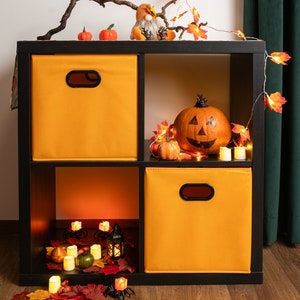 Puede incluir: Una estantería de cubos negros con cestas de almacenamiento de tela naranja. La repisa superior está decorada con una calabaza tallada, calabazas pequeñas, un gnomo y una rama con hojas de otoño y luces de cadena. La repisa inferior tiene una linterna y velas.