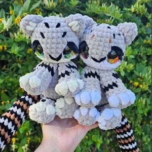 May include: Two handmade crochet tiger plush toys. The tigers are gray with black and white markings, and colorful eyes. They have white paws and striped tails. The toys are held in front of a green leafy background.