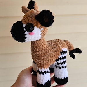 May include: A handmade plush okapi toy. The okapi has a brown body, black and white striped legs, black ears, and a black and white face. The toy has large black eyes and a pink mouth. The okapi is made of soft, textured yarn.