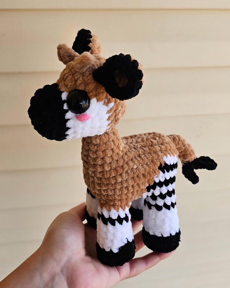 May include: A handmade plush okapi toy. The okapi has a brown body, black and white striped legs, black ears, and a black and white face. The toy has large black eyes and a pink mouth. The okapi is made of soft, textured yarn.