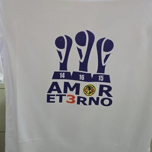 Puede incluir: Camiseta blanca con un gráfico azul y amarillo de tres trofeos con los números 14, 16 y 15 en ellos. El texto "AMOR ETERNO" está debajo de los trofeos.