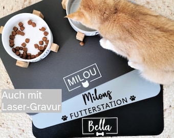 Puede incluir: Un gato come de un cuenco blanco sobre una alfombrilla de alimentación para mascotas negra y azul claro. La alfombrilla tiene el texto "MILOU" y "Milong Futterstation" con huellas de patas. El nombre "Bella" también está en la alfombrilla.
