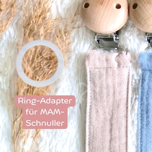 Könnte beinhalten: Nahaufnahme von Baby-Schnullerclips mit Holzperlen und Stoffbändern in Rosa und Blau. Das Bild enthält einen weißen Ringadapter und den Text "Ring-Adapter für MAM-Schnuller."