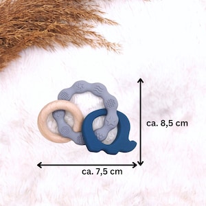 May include: A blue and gray silicone teether with a wooden ring. The teether is shaped like a letter Q and has a star and wave design. The teether measures approximately 7.5 inches wide and 8.5 inches tall.