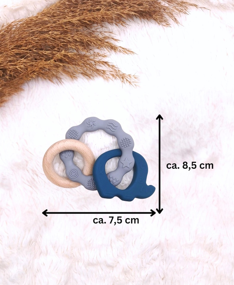 May include: A blue and gray silicone teether with a wooden ring. The teether is shaped like a letter Q and has a star and wave design. The teether measures approximately 7.5 inches wide and 8.5 inches tall.