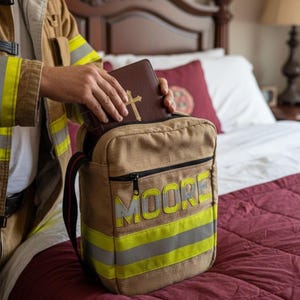 May include: A tan canvas gear bag featuring the name MOORE in yellow reflective lettering, accented with horizontal yellow and silver reflective safety stripes, and a black zipper closure, containing a brown book with a gold cross on the cover.