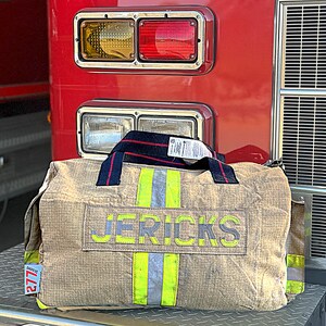 May include: A tan duffel bag with the name "JERICKS" in large, light green letters. The bag has reflective yellow and silver stripes and a dark blue and red striped handle. The bag is in front of a red vehicle with lights.