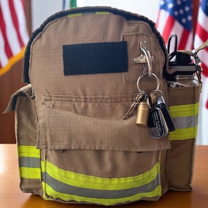 May include: A tan backpack with reflective yellow and gray stripes, a black patch, and a side pocket holding a water bottle. Keys and dog tags hang from a metal clip. The backpack is on a wooden surface with American flags in the background.