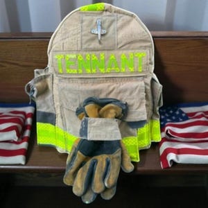 May include: A tan firefighter's coat and gloves are displayed, with the name "TENNANT" in bright green letters. The coat has a cross emblem and neon yellow trim. The gloves are tan and black. American flags are in the background.