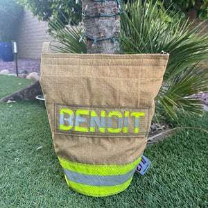 May include: A tan bucket-shaped bag with the name "BENOIT" in reflective yellow and gray letters. The bag has a reflective yellow and gray stripe around the bottom. The bag is made of a durable, possibly fire-resistant material.