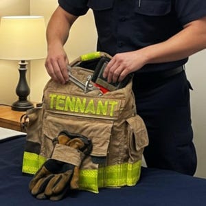 May include: Tan firefighter's gear bag with the name "TENNANT" in yellow lettering. The bag has multiple pockets and is open, revealing tools and a wallet. A pair of work gloves rests in front of the bag. The person is wearing a dark blue uniform.