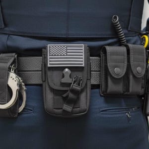 May include: A black tactical utility belt featuring a central pouch with a grey American flag patch, a side pouch for handcuffs, and a dual-compartment pouch with snap closures. The gear is attached to a textured black belt worn over dark blue trousers.