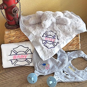 May include: A collection of baby items, including a white blanket with a firefighter emblem and the word "BABY" embroidered in pink. Also, a matching bib and pacifiers. The items are in shades of white and light blue.