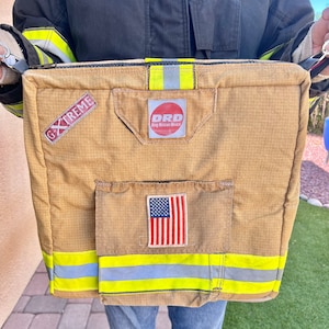 May include: Tan rectangular bag with the text "G XTREME" and "DRD Drag Rescue Device". Features a small American flag patch and reflective yellow stripes. The bag is held by a person wearing a dark jacket and blue jeans.