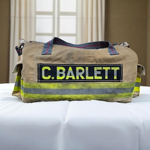 May include: A tan duffel bag with a black rectangular patch that reads "C. Bartlett" in bright yellow letters. The bag has a gray and yellow stripe design and a red and gray handle. The bag is sitting on a white surface.