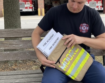 Op de afbeelding: Een rechthoekige, beige tas met een zwarte rits, een horizontale reflecterende gele streep en de naam MOORE in het geel gedrukt. Twee witte notitieblokken met spiraalbinding en de tekst FIRE ACADEMY zitten in het open hoofdvak.