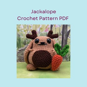 Könnte beinhalten: Ein gehäkeltes Jackalope mit großen schwarzen Augen, braunen Geweihen und einem dunkelbraunen Bauch. Es hält eine gehäkelte orangefarbene Karotte mit grünen Blättern. Der Text "Jackalope Crochet Pattern PDF" steht über dem Jackalope.
