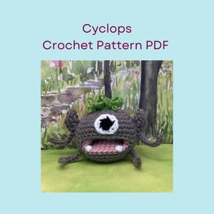 May include: A crocheted cyclops monster with one eye, grey body, and green hair. The monster has a wide mouth with pink and white details. The text "Cyclops Crochet Pattern PDF" is above the monster.