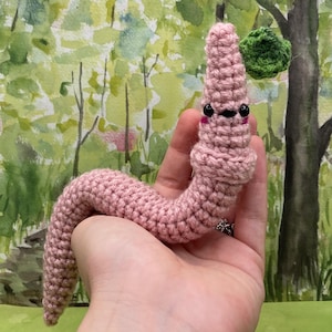 May include: A handmade, pink crochet worm with black eyes and a small mouth. The worm is held in a hand, with a green crocheted ball in the background. The worm is a whimsical, soft toy.