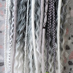 Puede incluir: Una colección de rastas grises, blancas y negras, algunas trenzadas, otras onduladas y otras con elementos decorativos. Las rastas cuelgan de una barra negra. Algunas rastas tienen encaje negro y cintas verde neón.